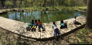 Six of our day off campers laying on the dock by the pond, gazing into the water, on a clear spring day