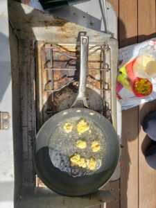 Yellow dandelion fritters frying in a pan of oil on a gas-powered camping stove, pictured from above
