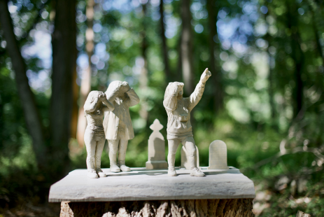 A sculpture of three small white figures with binoculars and small gravestones in the background mounted on a wooden stump in the forest.